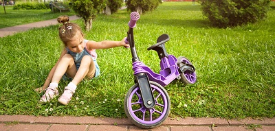 Fille joyeuse assise et se reposant sur l'herbe dans un parc près d'un vélo violet en été. Divertissement actif des enfants, scooter pour les tout-petits, enfant heureux. Espace de copie. Fond d'été